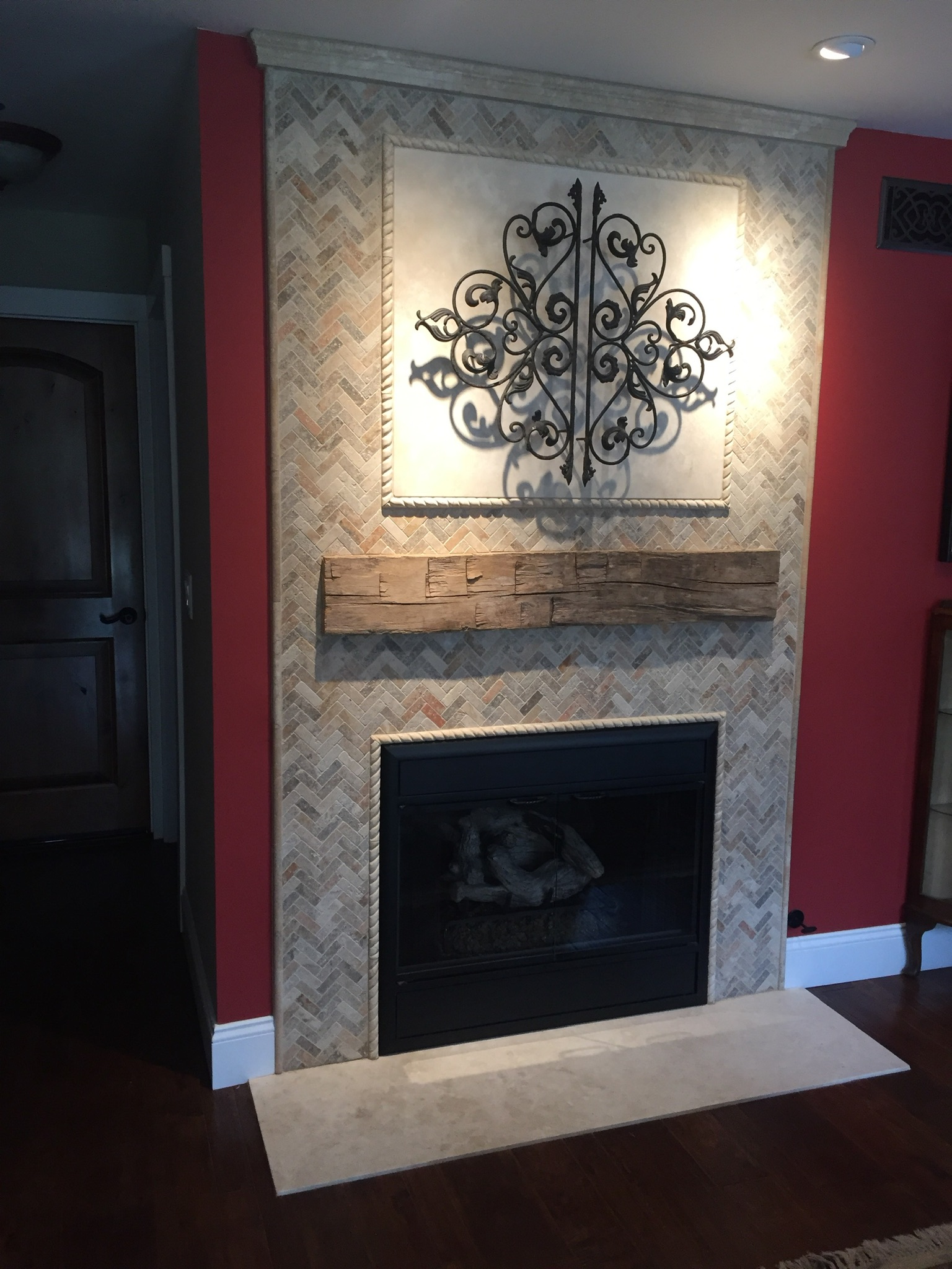 Strategically placed lights draw attention to an artistic design that hangs over a gorgeous backdrop, that is, a fireplace surround of multicolor tumbled travertine tiles set in a herringbone pattern.