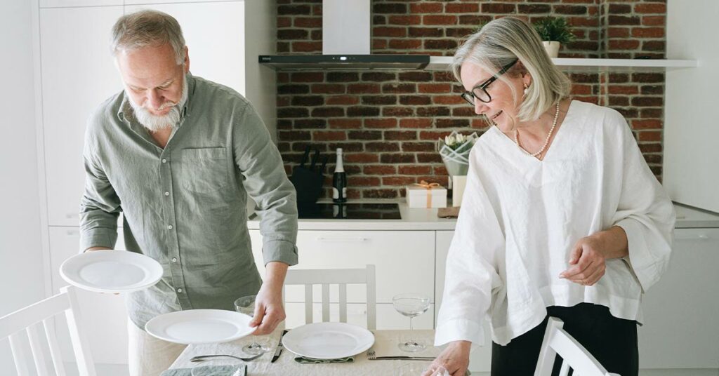 An older man and woman set the table in a brightly lit room with open shelves. They appear to be good candidates for an aging-in-place tile installation.