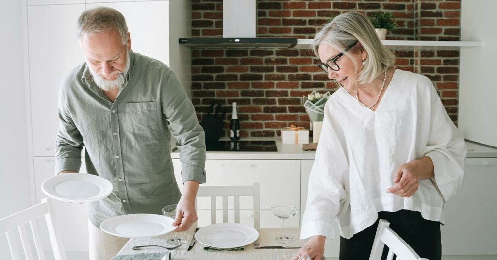 An older man and woman set the table in a brightly lit room with open shelves.