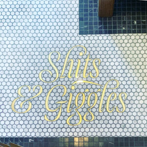 Hexagon marble bathroom tile floor has a funny custom brass inlay that says, "Shits & Giggles."